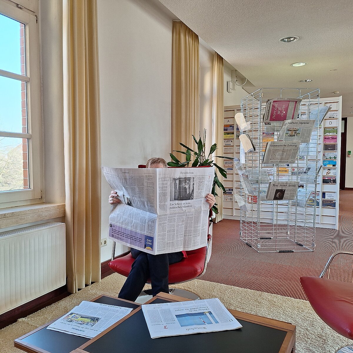 Eine Nutzerin sitzt in einem gemütlichen Drehsessel mit rotem Kunstlederpolster. Ihr Oberkörper ist nicht zu erkennen, da sie eine Zeitung aufgeschlagen vor sich hält. Im Vordergrung Zeitungen auf einem Tisch. Im Hintergrund ein Zeitungsständer und eine alphabetische Zeitschriftenablage.
