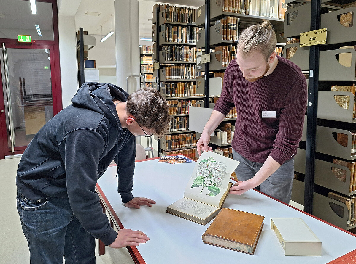 Ein Mitarbeiter zeigt einem Schüler eine Zeichnung in einem historischen Buch. Im Hintergrund Regalreihen mit alten Büchern in Leder- und Pergamenteinbänden.