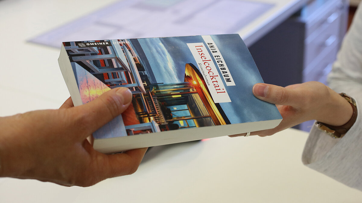 Eine Hand überreicht einer anderen ein Buch: "Inselcocktail. Kriminalroman" von Anja Eichbaum. Auf dem Cover Sitzgelegenheiten an Bord eines Schiffes oder an einem Pier in der Dämmerung.