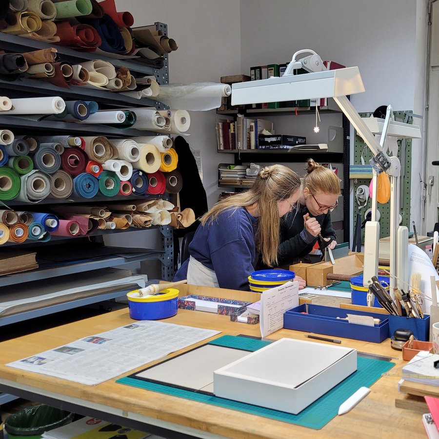 Werkstatt der Buchbinderei: Eine Buchbinderin in Ausbildung schaut ihrer Ausbilderin zu.