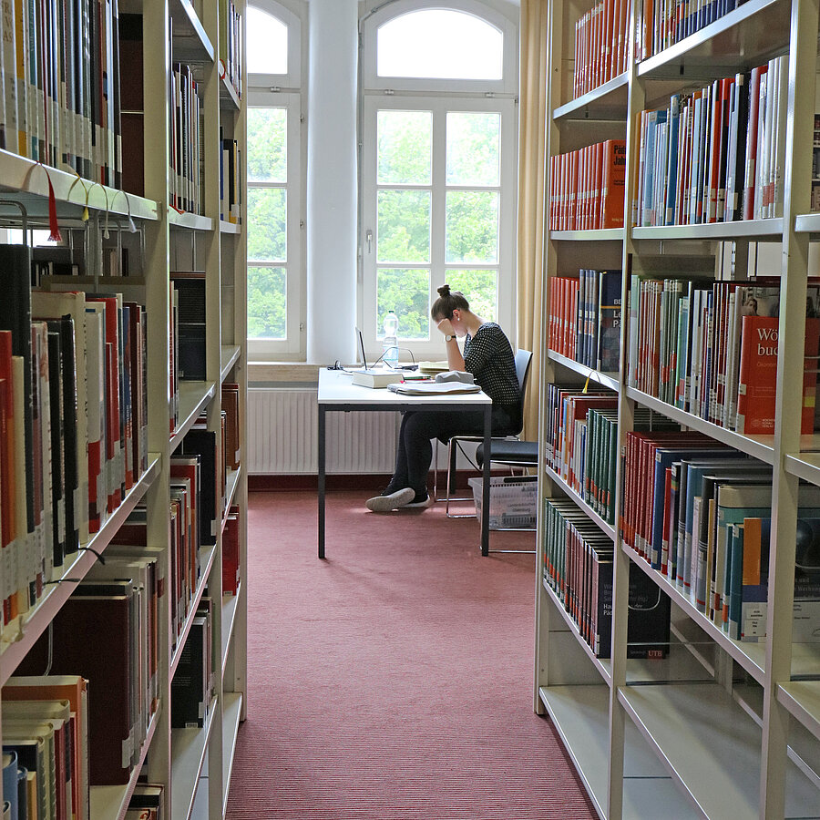 Blick durch zwei Regalreihen auf eine Nutzerin, die an einem Tisch arbeitet.