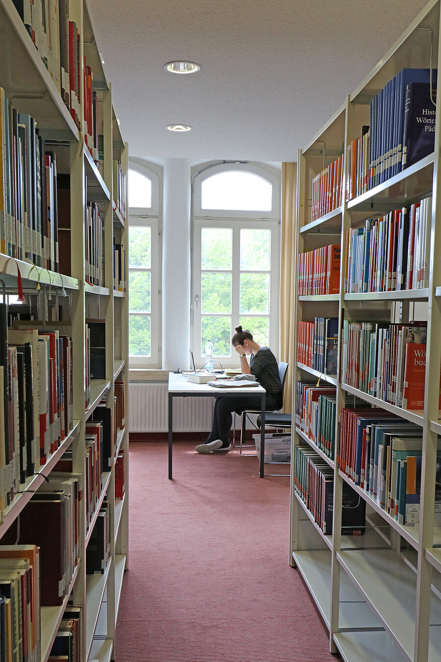 Blick durch einen Gang zwischen zwei Regalen. Im Fokus des Bildes eine Nutzerin, die an einem Tisch sitzt und liest.