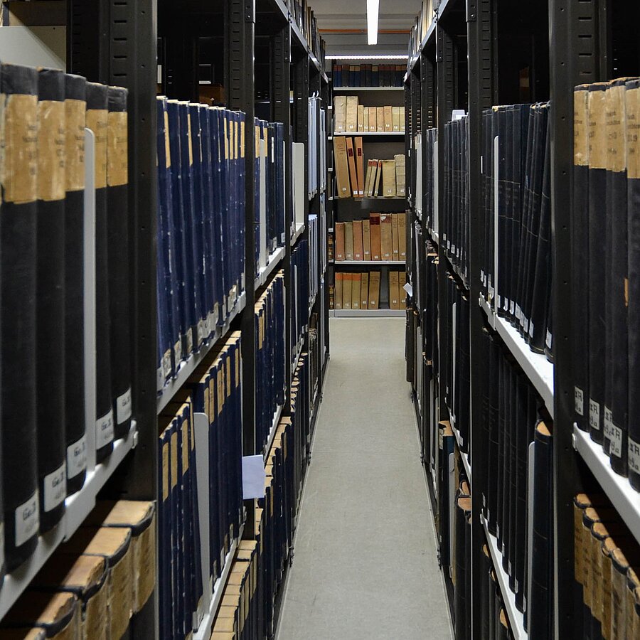 Blick durch einen Gang zwischen zwei Regalreihen mit Zeitungsbänden.