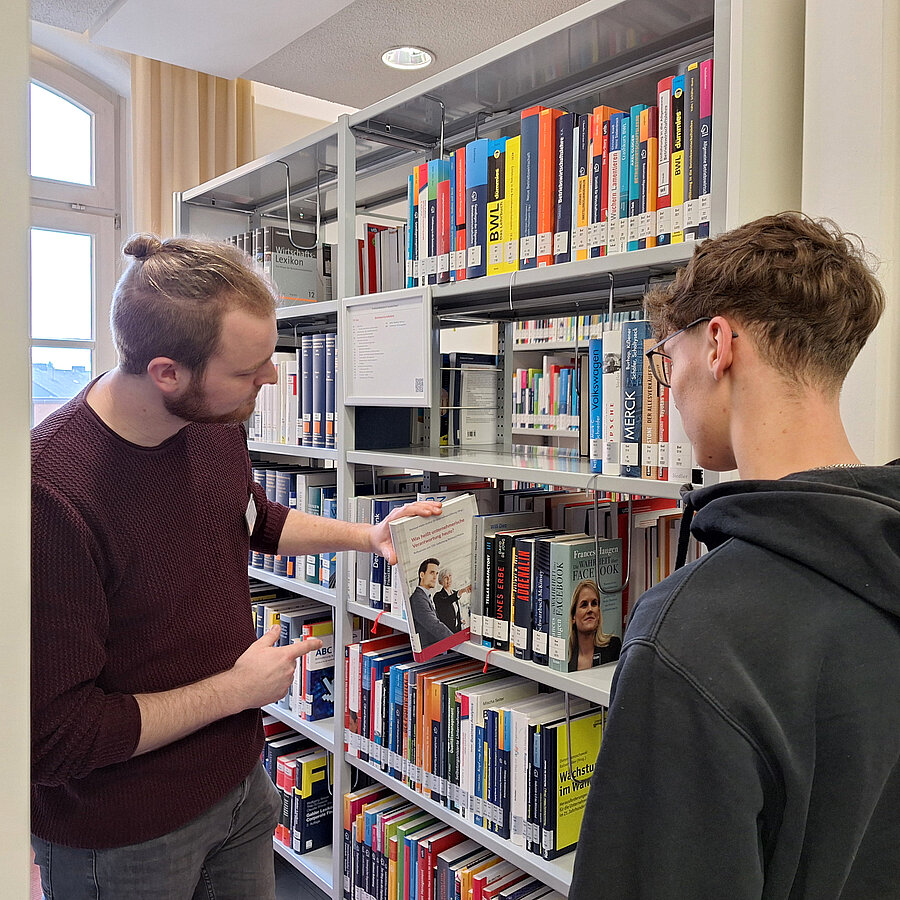 Ein Mitarbeiter zeigt einem Schüler ein Buch im Bücherregal.