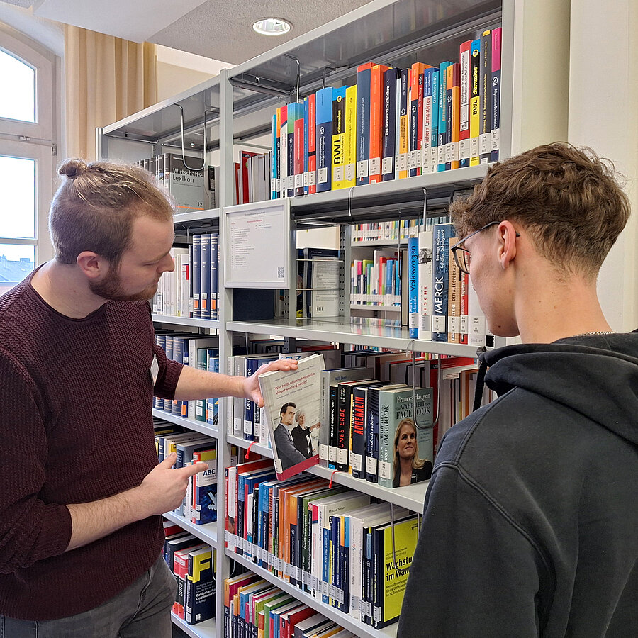 Ein Mitarbeiter zeigt einem Schüler am Regal ein Buch.