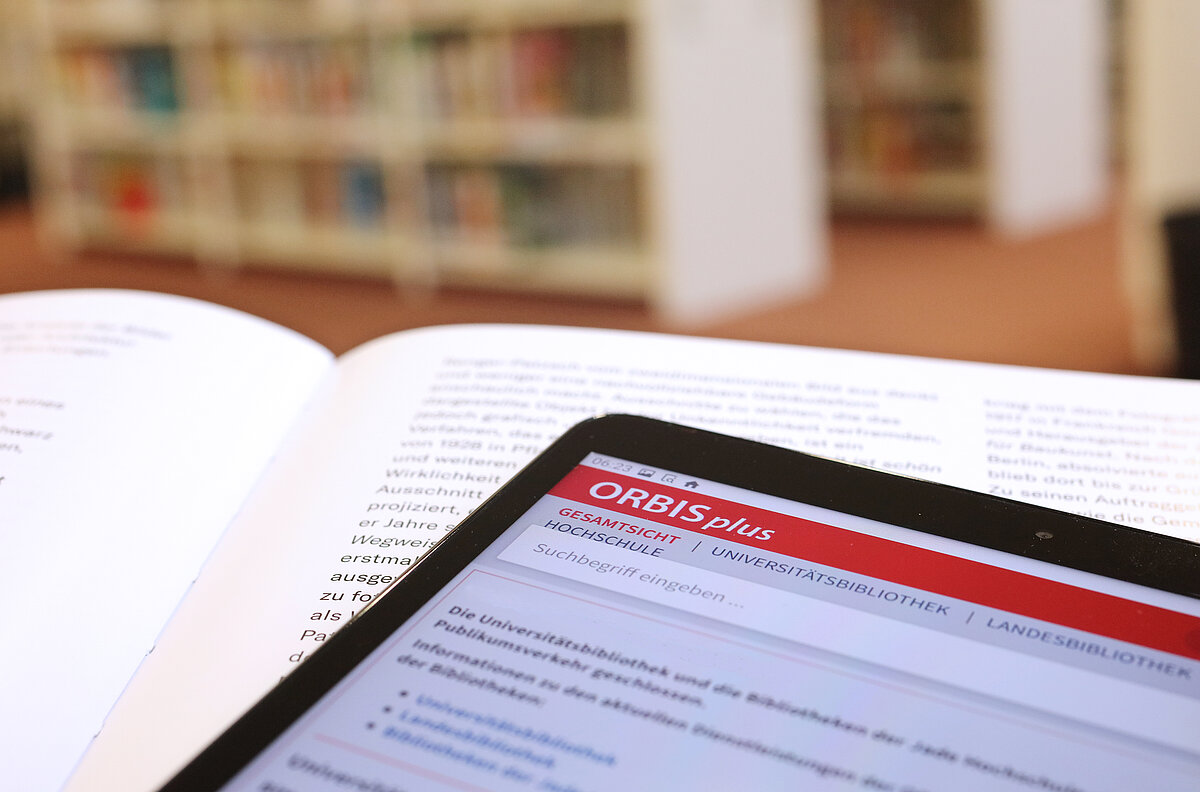 Ein Tablet mit dem Online-Katalog ORBISplus liegt auf einem aufgeschlagenen Buch. Im Hintergrund Regalreihen.