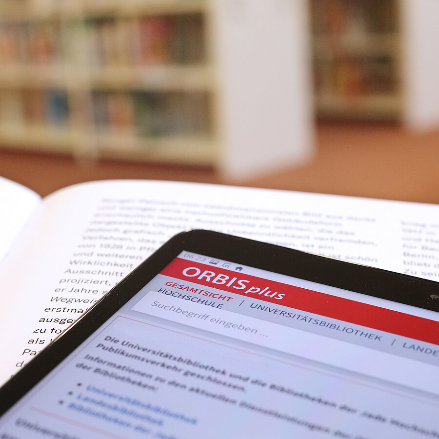 Ein Tablet liegt auf einem Buch und zeigt den Online-Katalog ORBISplus. Im Hintergrund Regalreihen.