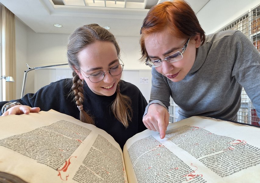 Nahaufnahme: Zwei Schüler:innen schauen in ein historisches Buch. Eine Schülerin zeigt mit dem Finger auf eine Textstelle.