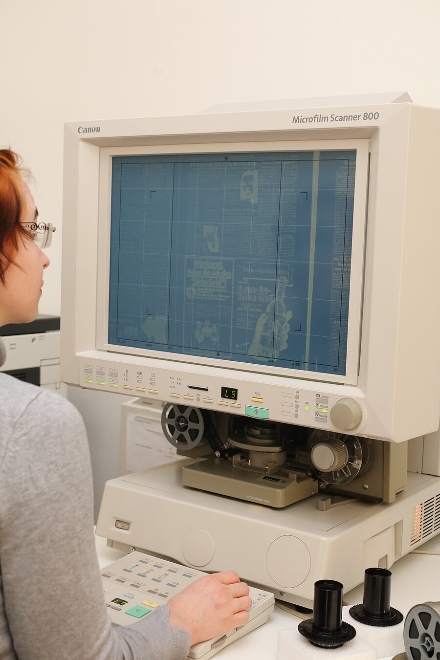 Eine Nutzerin sitzt vor einem Mikroform-Readerprinter. Der Bildschirm zeigt eine Zeitungsausgabe im Negativ.