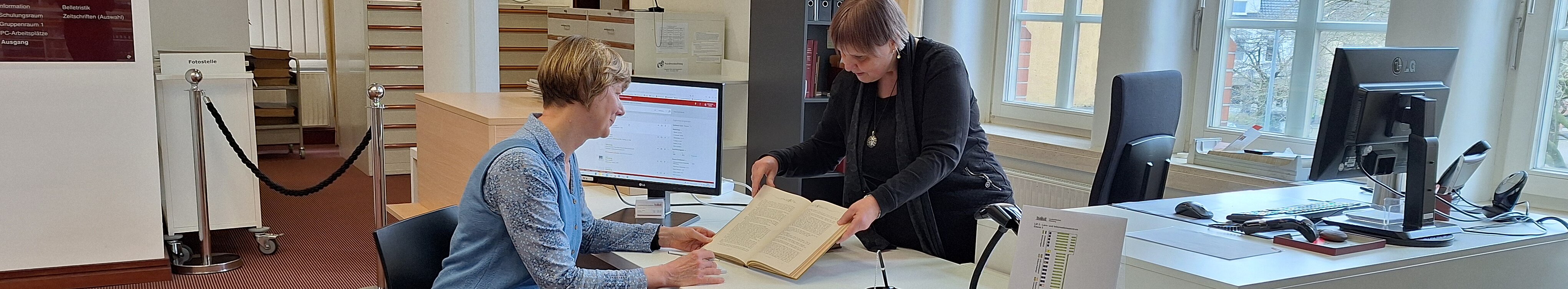 Eine Mitarbeiterin zeigt einer Nutzerin am Auskunftsplatz Ebene 2 (Lesesaal) ein historisches Buch.