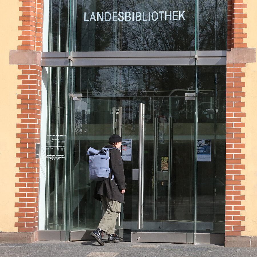 Eine Nutzerin mit schwarzer Jacke und Mütze sowie blauem Rucksack betritt den Haupteingang der Landesbibliothek Oldenburg.