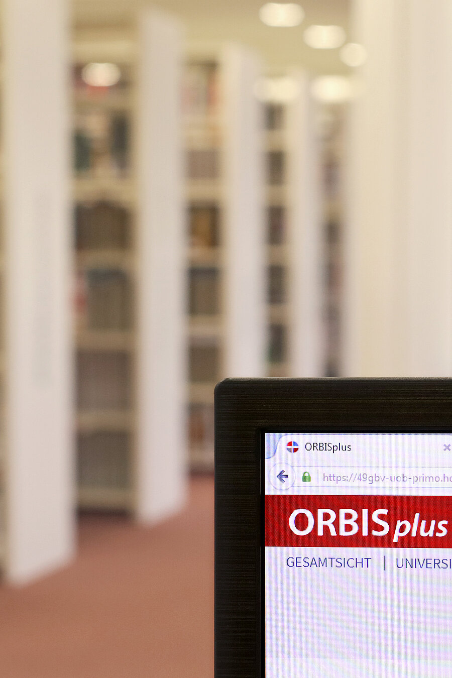 Ein Bildschirm zeigt den Online-Katalog ORBISplus. Im Hintergrund Regalreihen.