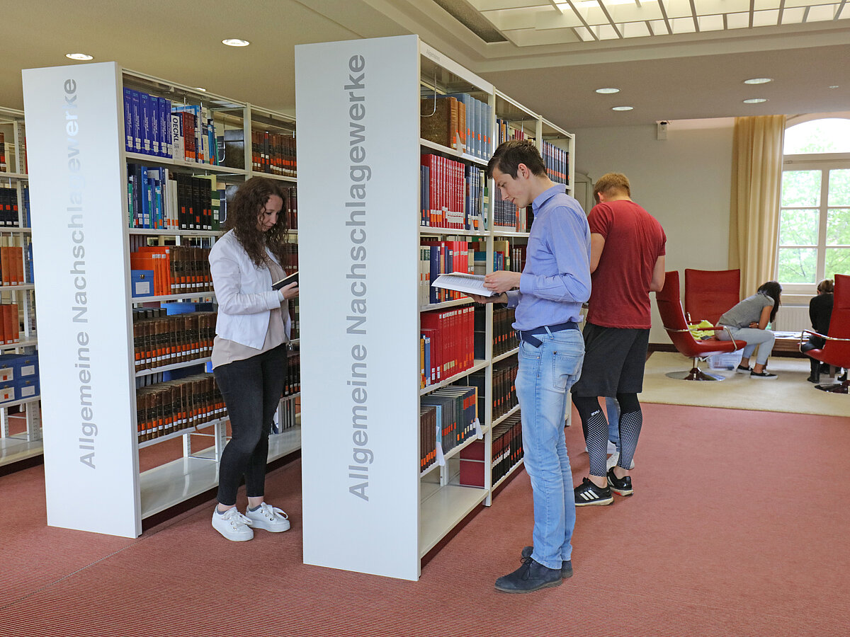 Drei Nutzer:innen stehen an zwei Regalen mit der Aufschrift "Allgemeine Nachschlagewerke" und blättern in Büchern.