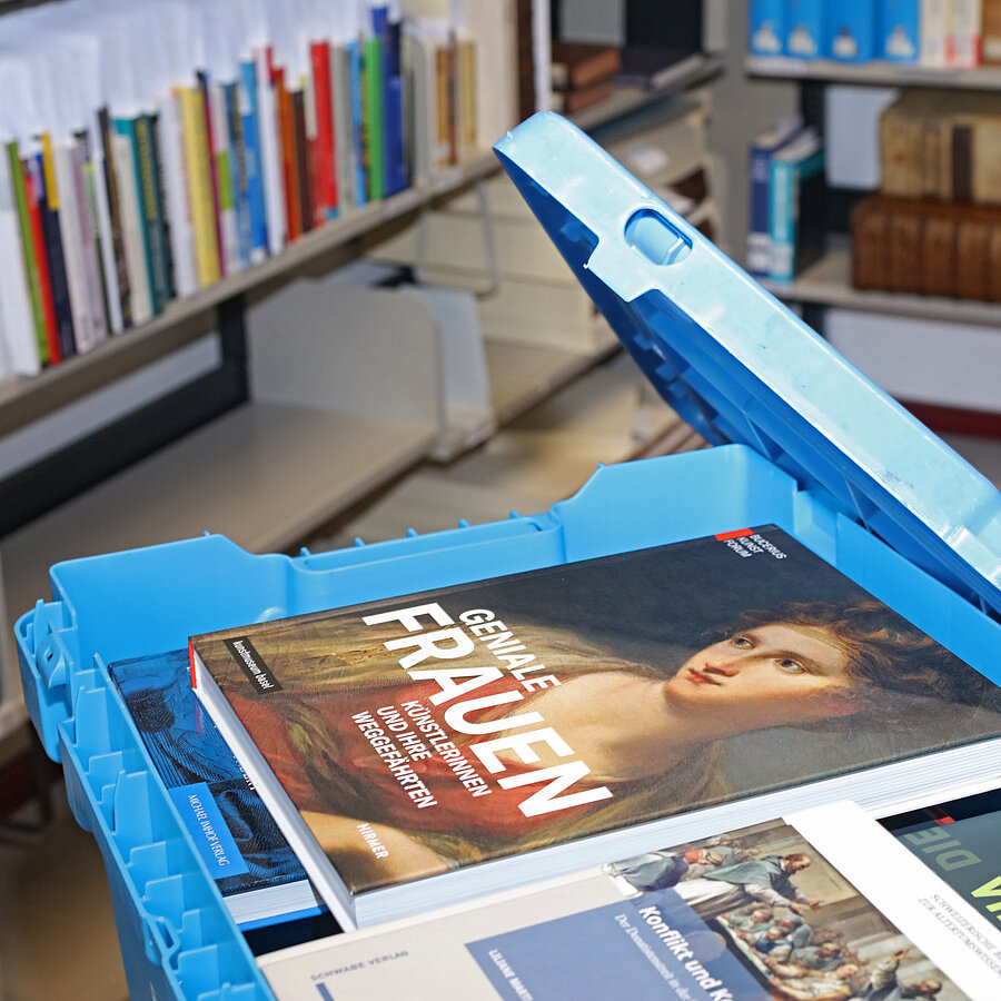 Blaue Bücherkiste mit geöffnetem Deckel. Fernleih-Bücher befinden sich in der Kiste. Im Hintergrund Bücher im Regal.