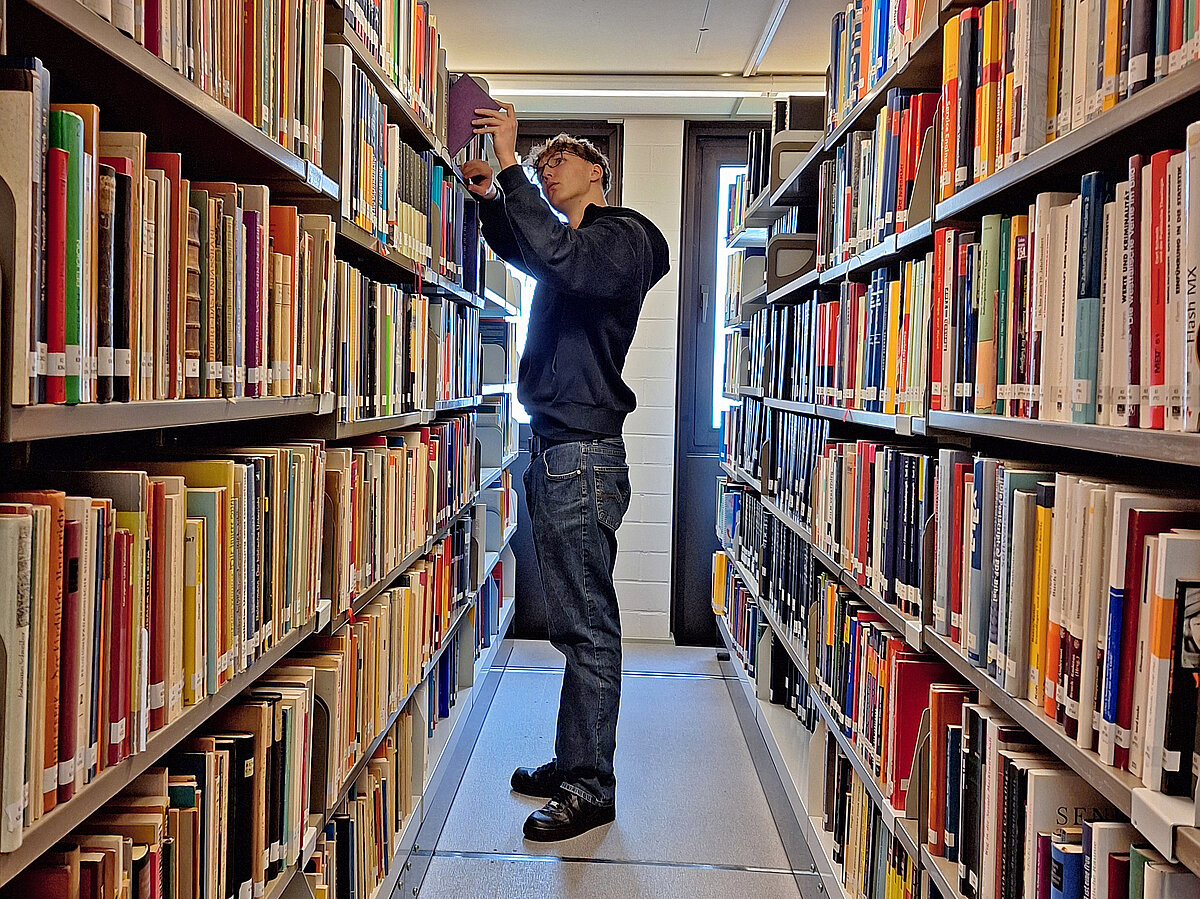 Ein Schüler steht in einem Gang zwischen zwei Bücherregalen und zieht ein Buch heraus.