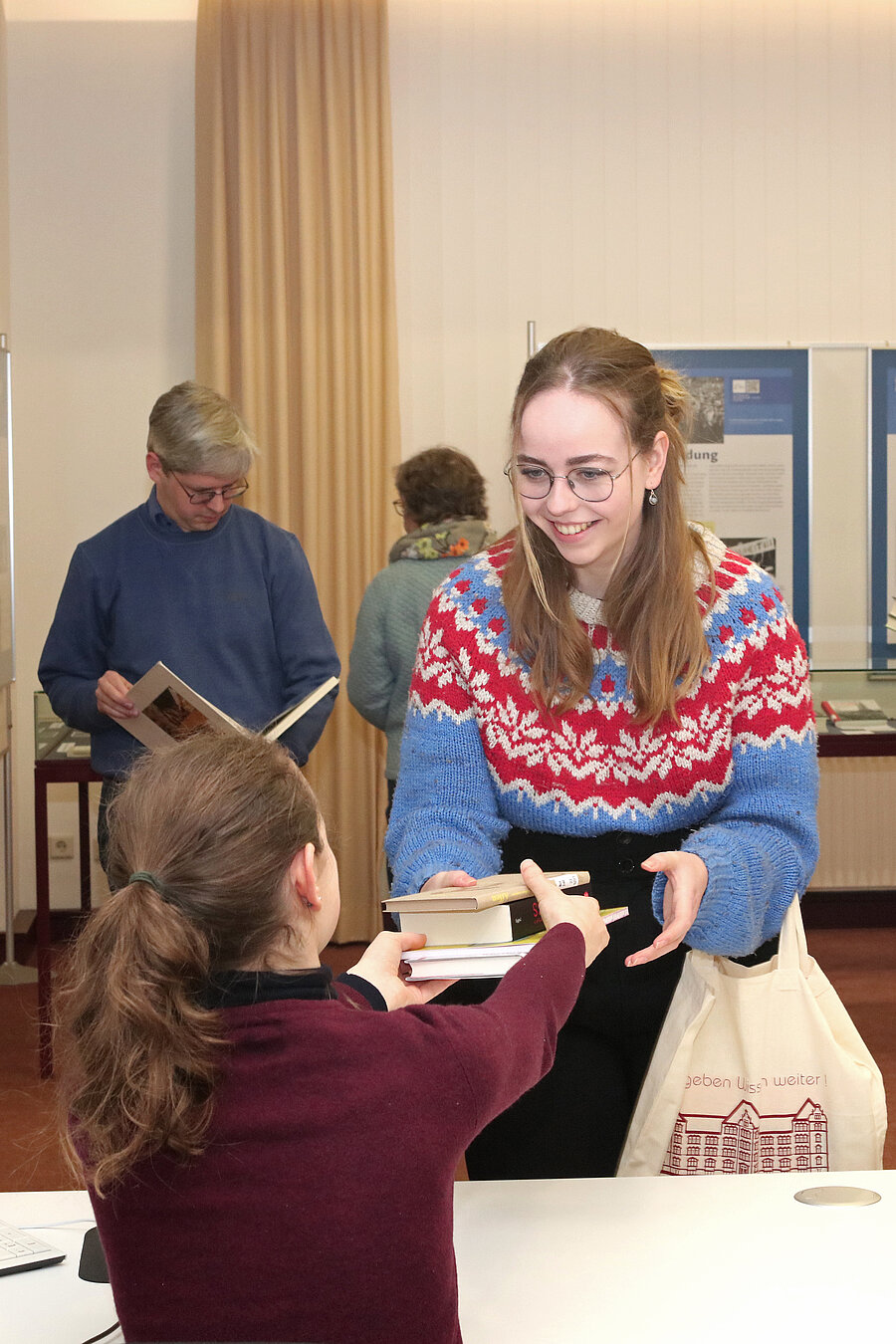 Blick über die Ausleihtheke und über den Rücken der Bibliotheksmitarbeiterin, wie sie einer Nutzerin Bücher aushändigt. Diese lächelt freundlich.Im Hintergrund wartende Nutzer:innen.